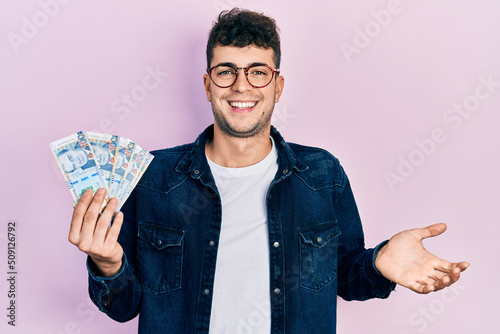 Young hispanic man holding peruvian sol banknotes celebrating achievement with happy smile and winner expression with raised hand