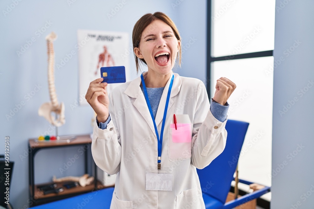 Young brunette woman working at pain recovery clinic holding credit card screaming proud, celebrating victory and success very excited with raised arms