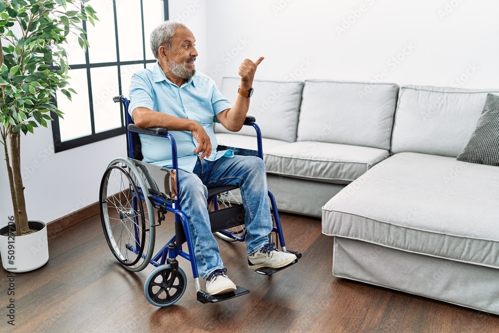 Handsome senior man sitting on wheelchair at the living room smiling with happy face looking and pointing to the side with thumb up. Handsome senior man sitting on wheelchair at the living room smiling with happy face looking and pointing to the side with thumb up.