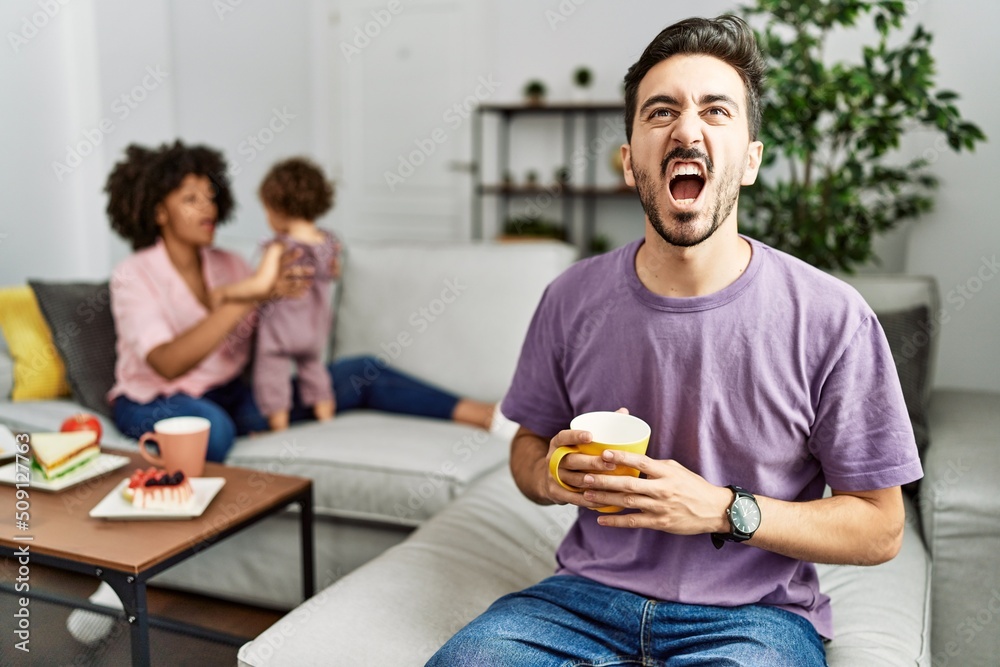 Hispanic father of interracial family drinking a cup coffee angry and mad screaming frustrated and furious, shouting with anger. rage and aggressive concept.