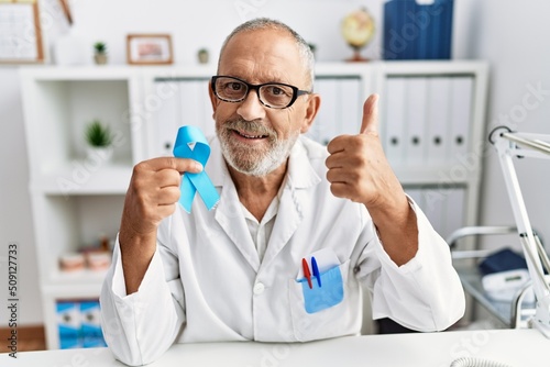 Mature doctor man holding blue ribbon at the clinic smiling happy and positive, thumb up doing excellent and approval sign