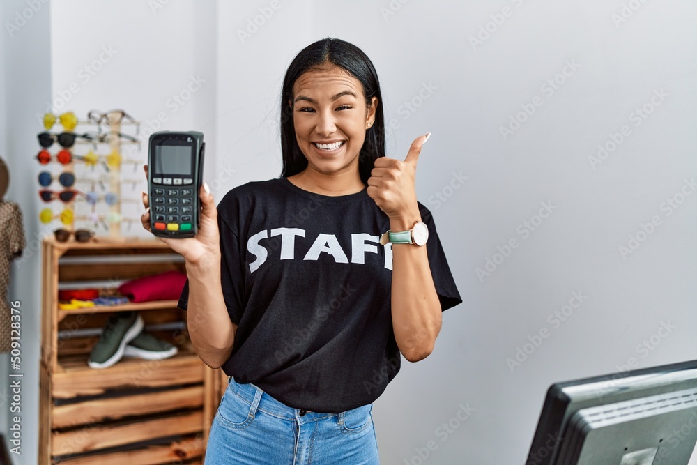 Young hispanic woman working at retail boutique holding dataphone smiling happy and positive, thumb up doing excellent and approval sign Young hispanic woman working at retail boutique holding dataphone smiling happy and positive, thumb up doing excellent and approval sign