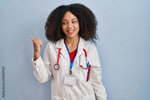 Young african american woman wearing doctor uniform and stethoscope smiling with happy face looking and pointing to the side with thumb up.