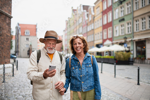 Portrait of happy senior couple tourists smiling, holding hands, using smartphone outdoors in historic town © Halfpoint