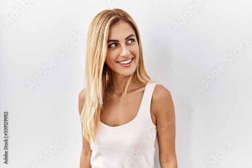 Blonde beautiful young woman standing over white isolated background looking away to side with smile on face, natural expression. laughing confident.