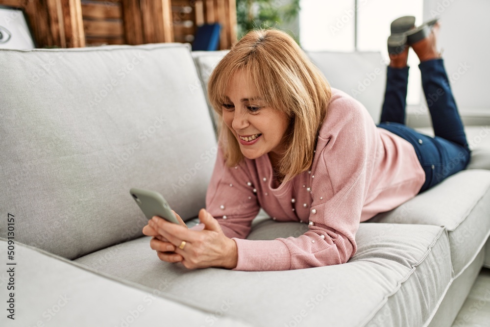 Obraz premium Middle age blonde woman using smartphone lying on the sofa at home.