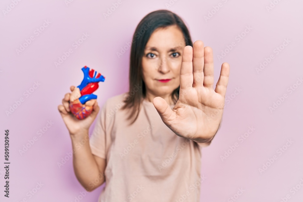 Middle age hispanic woman holding heart organ with open hand doing stop sign with serious and confident expression, defense gesture Middle age hispanic woman holding heart organ with open hand doing stop sign with serious and confident expression, defense gesture