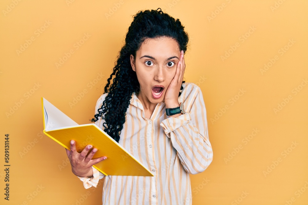 Young hispanic woman with curly hair holding book afraid and shocked, surprise and amazed expression with hands on face Young hispanic woman with curly hair holding book afraid and shocked, surprise and amazed expression with hands on face