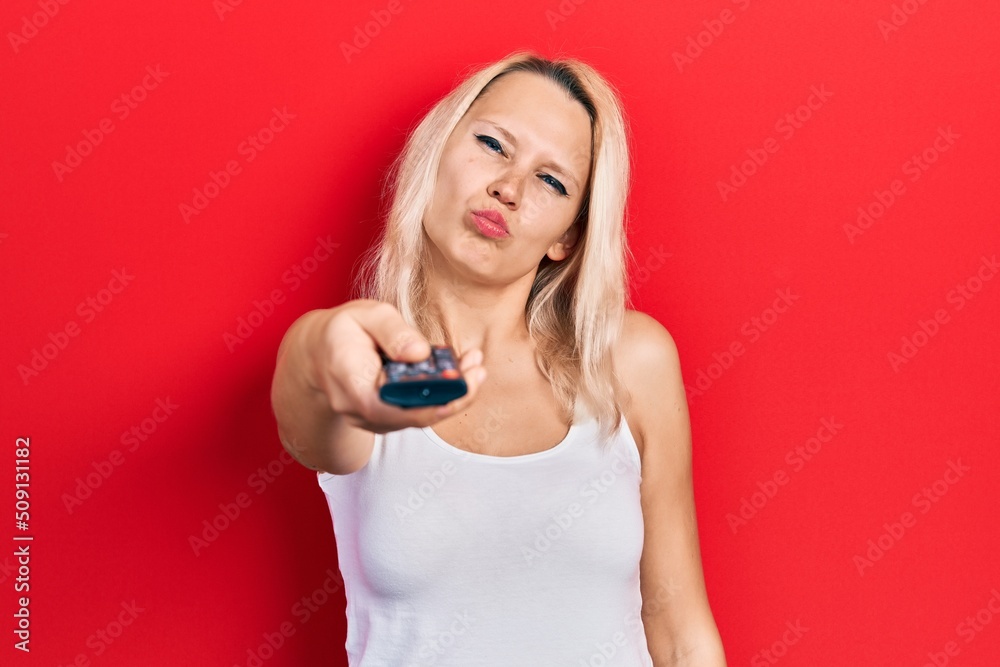 Beautiful caucasian blonde woman holding television remote control looking at the camera blowing a kiss being lovely and sexy. love expression. Beautiful caucasian blonde woman holding television remote control looking at the camera blowing a kiss being lovely and sexy. love expression.