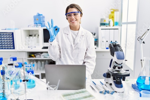 Young hispanic woman wearing scientist uniform working at laboratory doing ok sign with fingers  smiling friendly gesturing excellent symbol