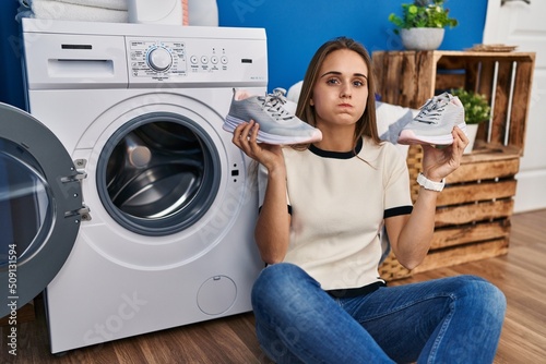 Young woman putting sneakers in washing machine puffing cheeks with funny face. mouth inflated with air, catching air.