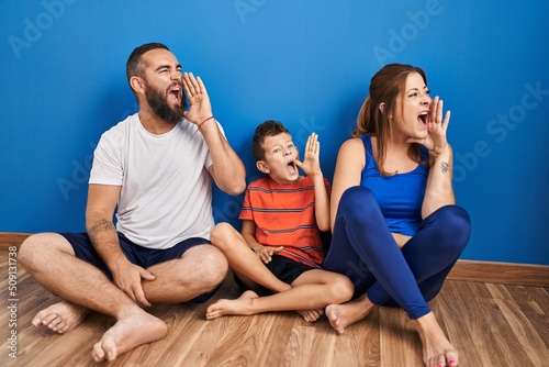 Family of three sitting on the floor at home shouting and screaming loud to side with hand on mouth. communication concept.