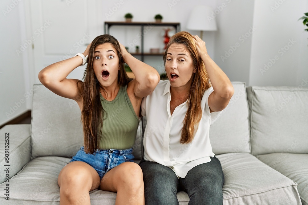 Mother and daughter together sitting on the sofa at home crazy and scared with hands on head, afraid and surprised of shock with open mouth Mother and daughter together sitting on the sofa at home crazy and scared with hands on head, afraid and surprised of shock with open mouth