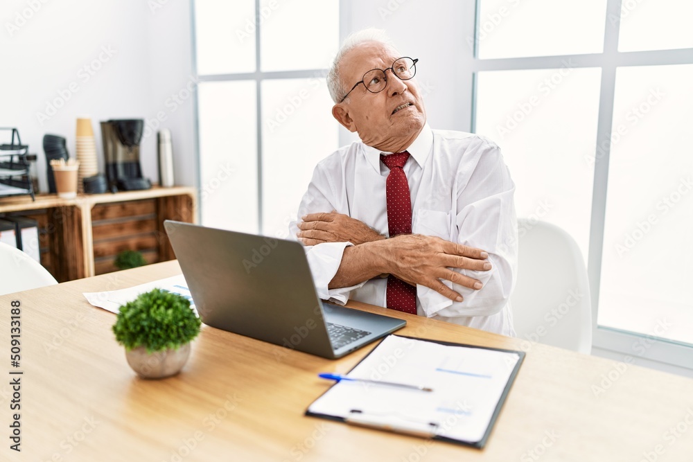 Senior man working at the office using computer laptop looking to the side with arms crossed convinced and confident Senior man working at the office using computer laptop looking to the side with arms crossed convinced and confident
