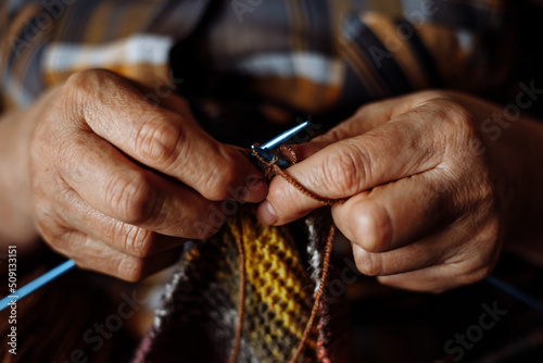 Close-up of hands of unrecognizable senior woman wearing checkered shirt, knitting colorful scarf with metal needles.