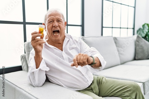 Senior man holding pills angry and mad screaming frustrated and furious, shouting with anger. rage and aggressive concept.