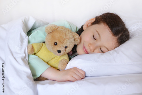 Beautiful cute little girl with a toy teddy bear sleeps in her bed, smiling sweetly