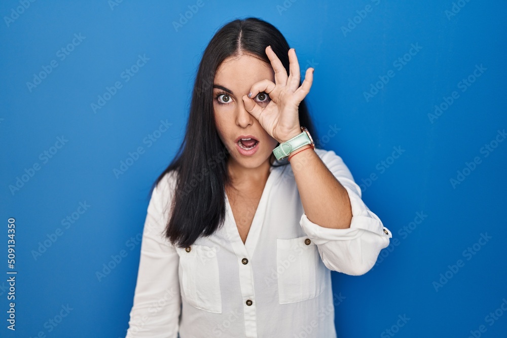 Young hispanic woman standing over blue background doing ok gesture shocked with surprised face, eye looking through fingers. unbelieving expression. Young hispanic woman standing over blue background doing ok gesture shocked with surprised face, eye looking through fingers. unbelieving expression.