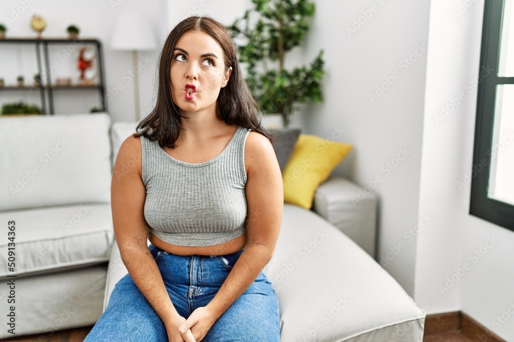 Young brunette woman sitting on the sofa at home making fish face with lips, crazy and comical gesture. funny expression. Young brunette woman sitting on the sofa at home making fish face with lips, crazy and comical gesture. funny expression.