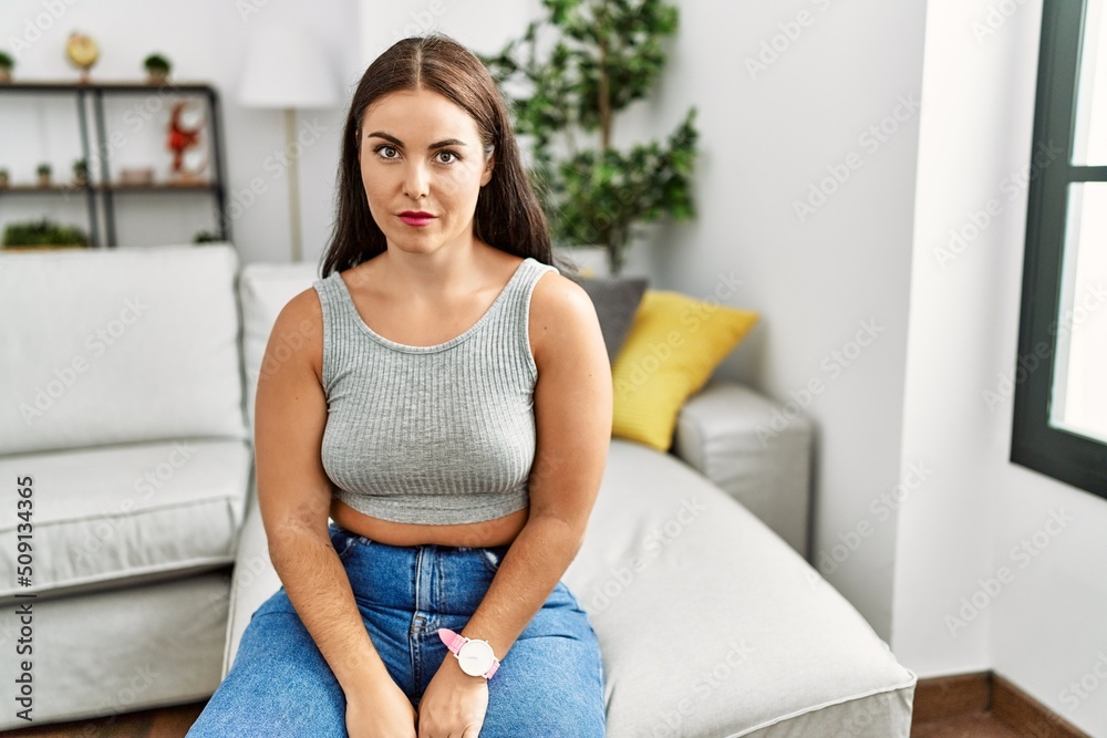 Young brunette woman sitting on the sofa at home relaxed with serious expression on face. simple and natural looking at the camera. Young brunette woman sitting on the sofa at home relaxed with serious expression on face. simple and natural looking at the camera.