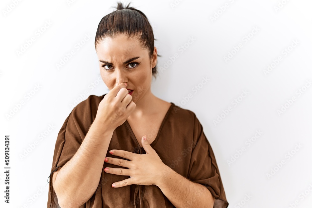 Young brunette woman standing over isolated background smelling something stinky and disgusting, intolerable smell, holding breath with fingers on nose. bad smell Young brunette woman standing over isolated background smelling something stinky and disgusting, intolerable smell, holding breath with fingers on nose. bad smell