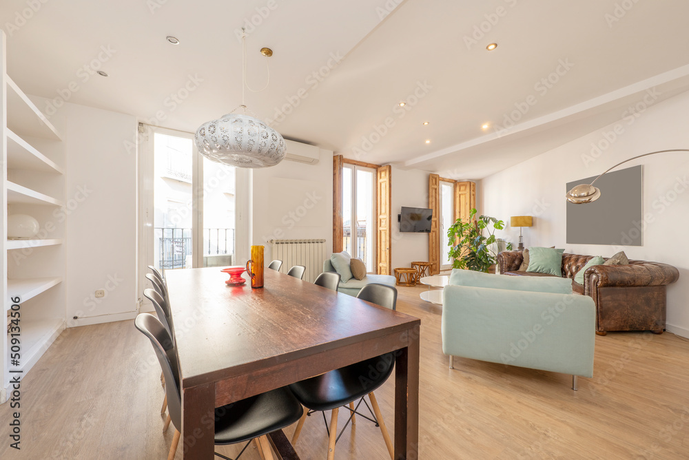 Fototapeta premium Living room with large wooden dining table and black resin chairs, three-seater chester sofa, plaster work shelving and wooden floor