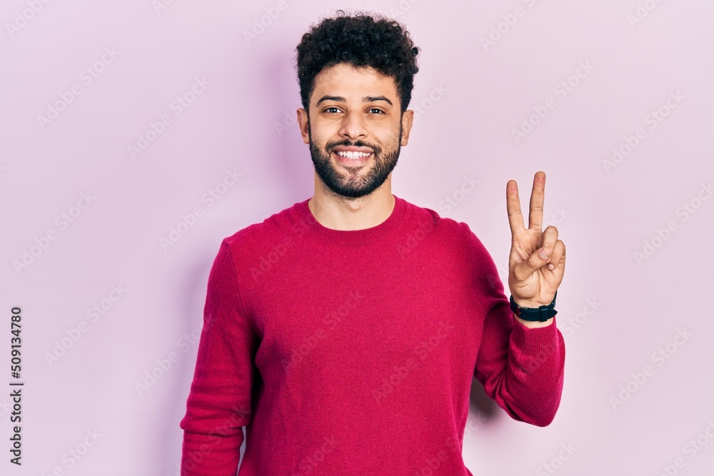 Young arab man with beard wearing casual pink sweater smiling with happy face winking at the camera doing victory sign. number two.