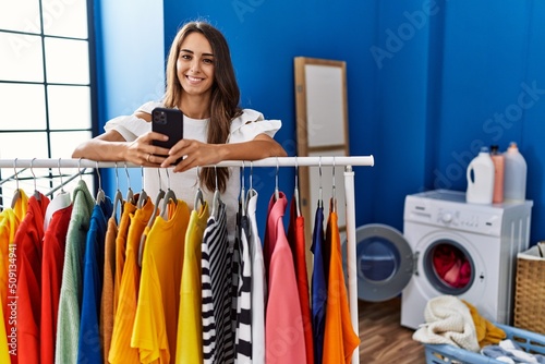 Young hispanic woman using smartphone leaning on rack at laundry room