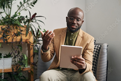 Young confident Afrcan American male psychologist with digital tablet in hand consulting patient during online session in his office photo