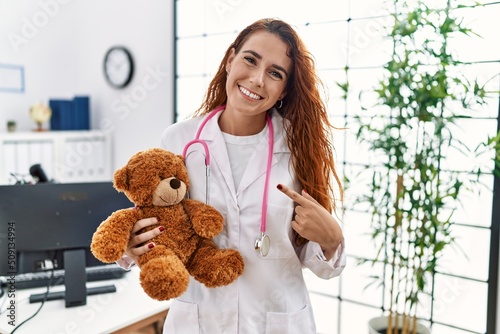 Young redhead pedriatician woman holding teddy bear smiling happy pointing with hand and finger photo