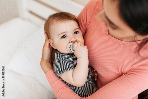 Loving mother holding cute infant baby in arms, little boy or girl with pacifier in mouth looking at camera, copy space