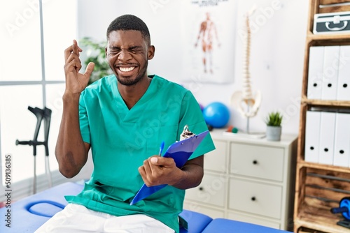 Young african american man working at pain recovery clinic gesturing finger crossed smiling with hope and eyes closed. luck and superstitious concept.