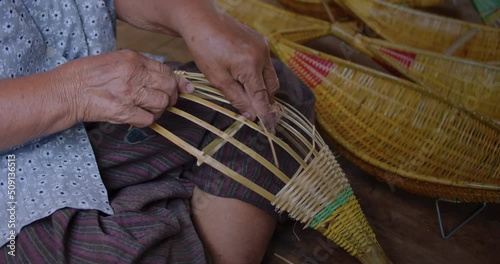 Cut in shot of an Asian female farmer who is an elderly native artisan weave souvenir container in the shape of a small cradle from bamboo that are cut into strips for weaving folk crafts.