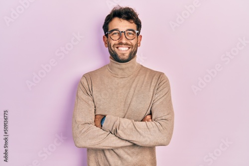 Handsome man with beard wearing turtleneck sweater and glasses happy face smiling with crossed arms looking at the camera. positive person.