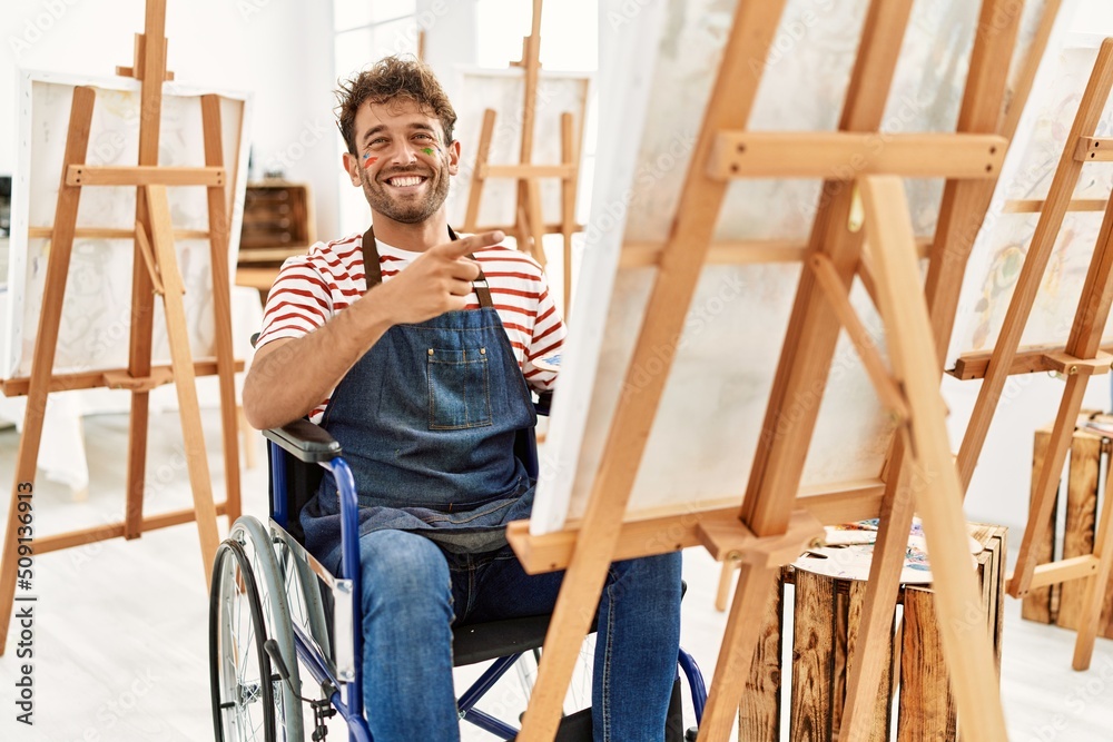 Young handsome man with beard at art studio sitting on wheelchair smiling happy pointing with hand and finger