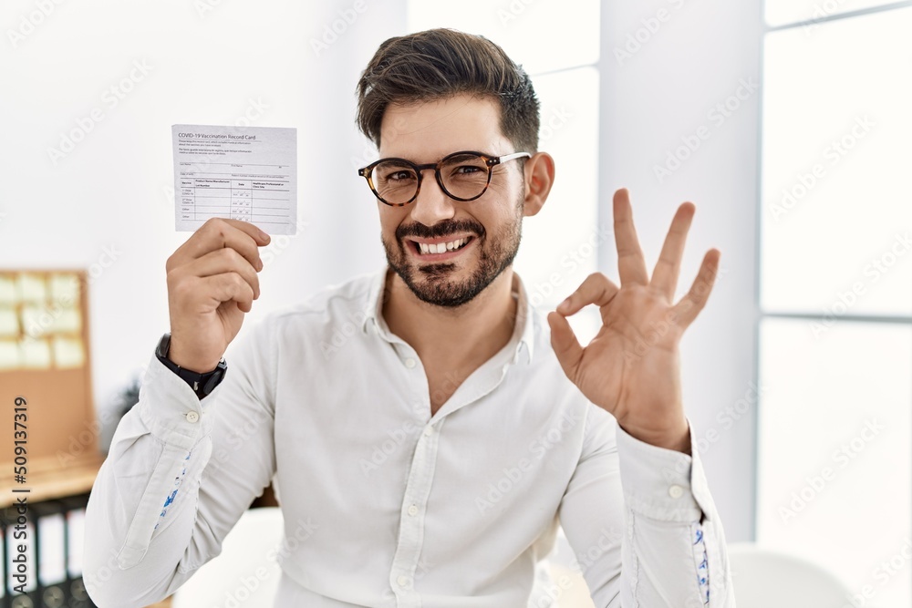 Young man with beard holding covid record card doing ok sign with fingers, smiling friendly gesturing excellent symbol Young man with beard holding covid record card doing ok sign with fingers, smiling friendly gesturing excellent symbol