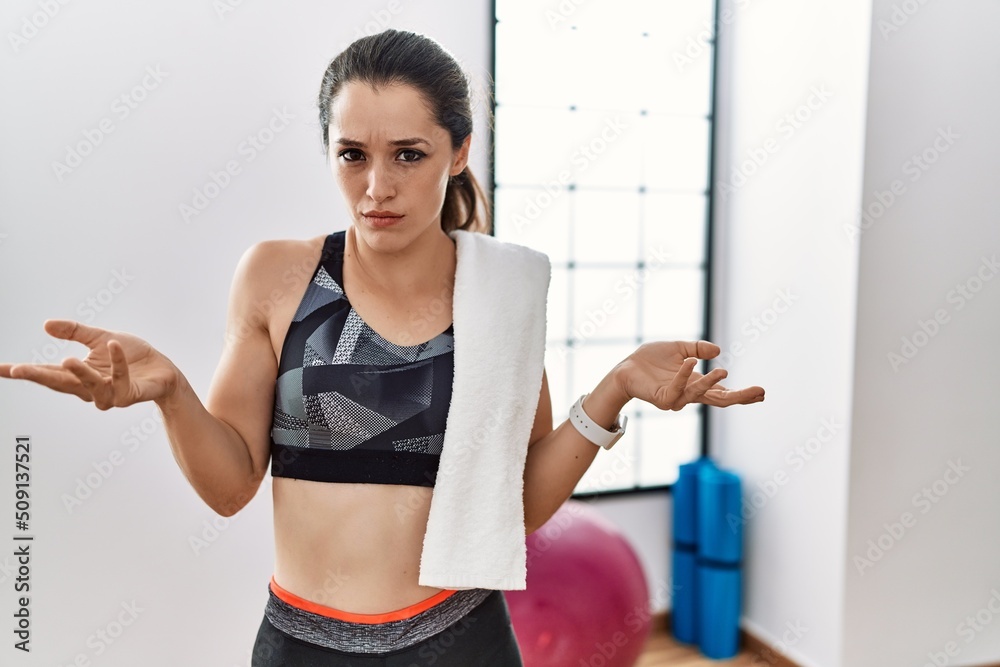 Young brunette woman wearing sportswear and towel at the gym clueless and confused with open arms, no idea concept.