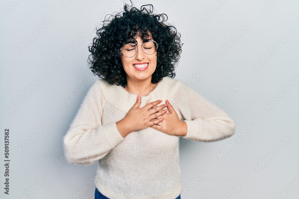 Young middle east woman wearing casual white tshirt smiling with hands on chest with closed eyes and grateful gesture on face. health concept. Young middle east woman wearing casual white tshirt smiling with hands on chest with closed eyes and grateful gesture on face. health concept.
