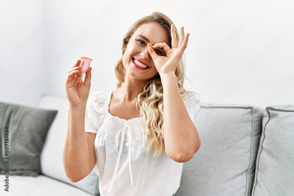 Blonde young woman holding menstrual cup smiling happy doing ok sign with hand on eye looking through fingers Blonde young woman holding menstrual cup smiling happy doing ok sign with hand on eye looking through fingers