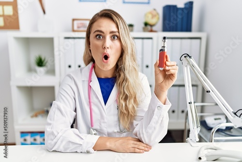 Young beautiful doctor woman holding electronic cigarette at the clinic scared and amazed with open mouth for surprise, disbelief face