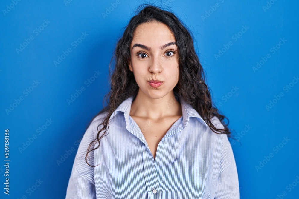 Young brunette woman standing over blue background puffing cheeks with funny face. mouth inflated with air, crazy expression. Young brunette woman standing over blue background puffing cheeks with funny face. mouth inflated with air, crazy expression.