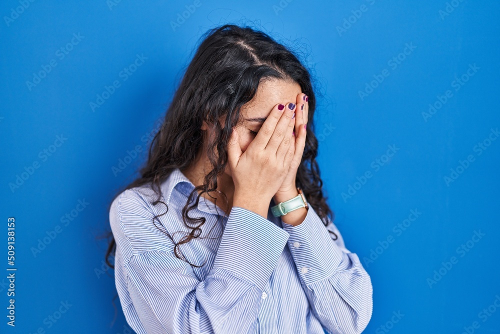 Young brunette woman standing over blue background with sad expression covering face with hands while crying. depression concept. Young brunette woman standing over blue background with sad expression covering face with hands while crying. depression concept.