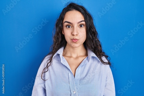 Young brunette woman standing over blue background puffing cheeks with funny face. mouth inflated with air, crazy expression.