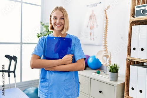Young blonde woman wearing physiotherapist uniform holding clipboard at rehab clinic