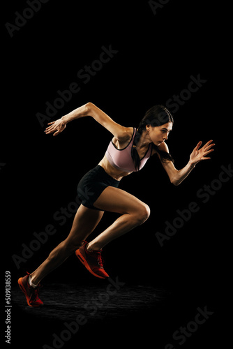 Studio shot of young muscular woman running isolated on black background. Sport, track-and-field athletics, competition and active lifestyle concept