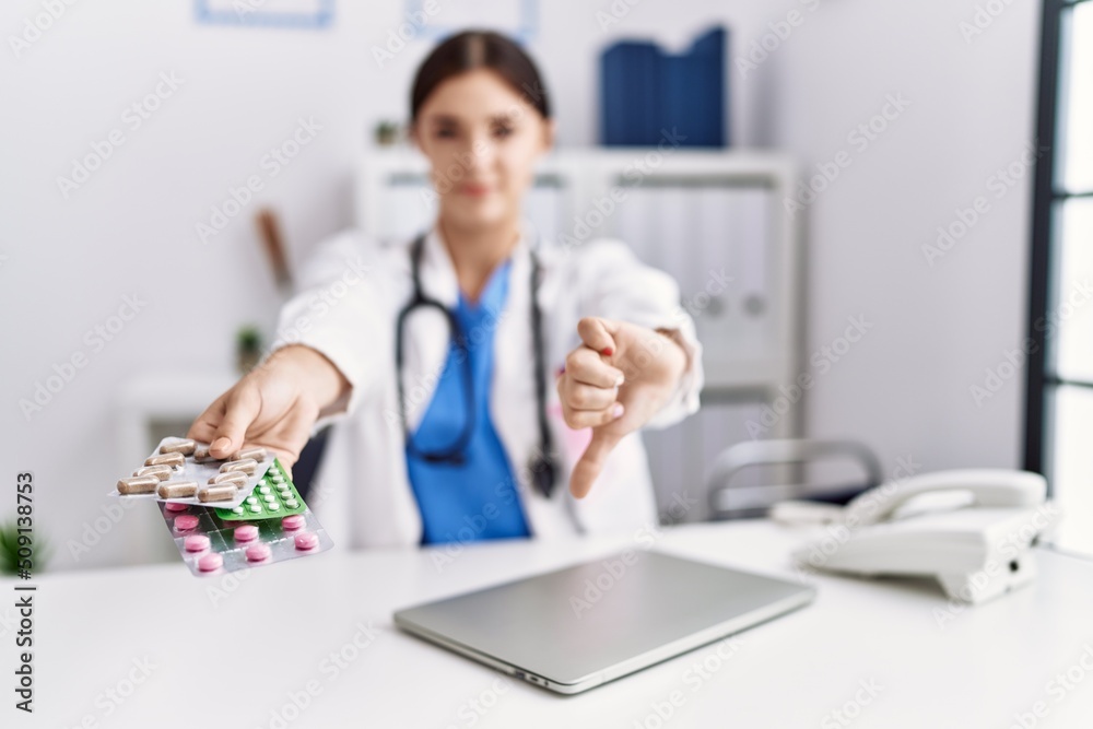 Young hispanic doctor woman wearing doctor uniform holding prescription pills at clinic with angry face, negative sign showing dislike with thumbs down, rejection concept Young hispanic doctor woman wearing doctor uniform holding prescription pills at clinic with angry face, negative sign showing dislike with thumbs down, rejection concept