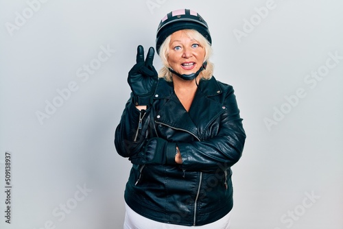 Middle age blonde woman holding motorcycle helmet smiling with happy face winking at the camera doing victory sign. number two.