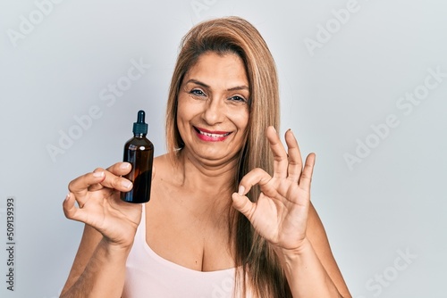 Middle age hispanic woman using night serum doing ok sign with fingers, smiling friendly gesturing excellent symbol