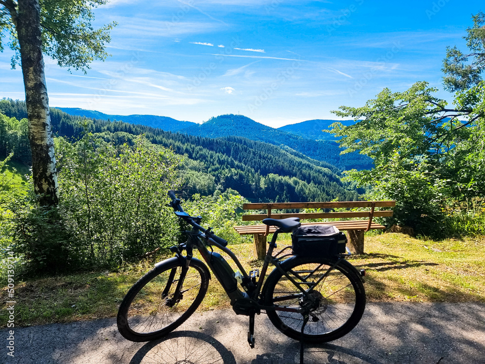 Fototapeta premium Electric Bicycle stands on a viewing platform in the beautiful countryside. Panoramic viewpoint with green vegetation, mountains and blue sky. Black Forest, Ortenau, Germany.