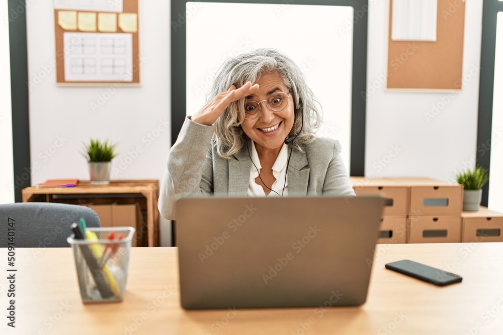 Middle age businesswoman sitting on desk working using laptop at office very happy and smiling looking far away with hand over head. searching concept.